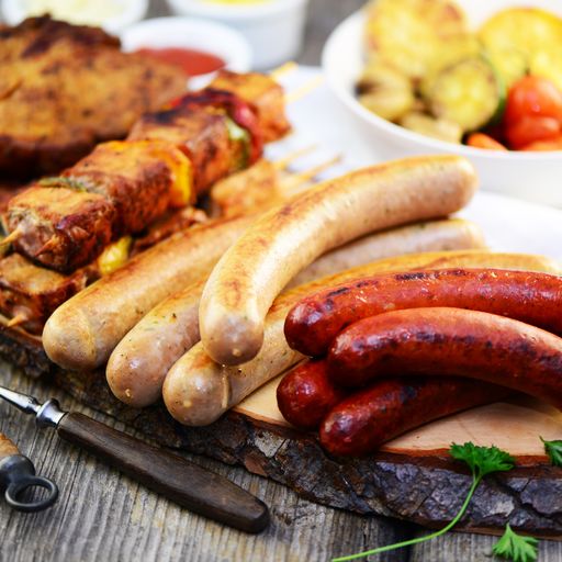 Zwei verschiedene Grillwürstchen und ein Fleischspieß auf einem Grill und dahinter eine weiße Schüssel mit Kartoffeln