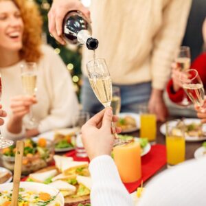 Neujahrs-Brunch mit fröhlicher Frau an gedeckter Tafel und vier Hände mit einem Glas Sekt, die sich zu prosten. Und eine Hand, die mit Flasche Sekt, die einschenkt einschenkt.