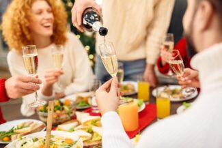 Neujahrs-Brunch mit fröhlicher Frau an gedeckter Tafel und vier Hände mit einem Glas Sekt, die sich zu prosten. Und eine Hand, die mit Flasche Sekt, die einschenkt einschenkt.