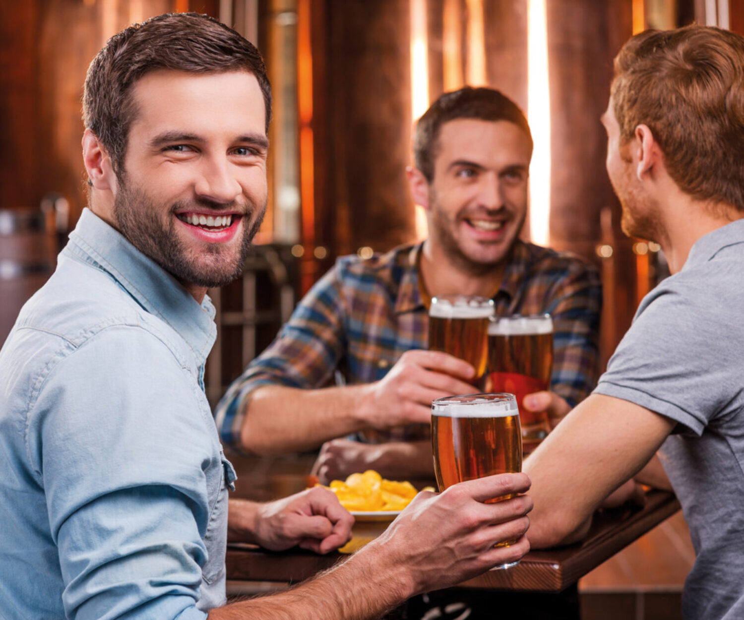 Spending time with best friends. Handsome young man toasting with beer and smiling while sitting with his friends in beer pub