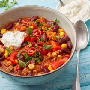 Hellblaue Bowlschale mit Chili sin Carne. Rechts davon Eßlöffel angelegt